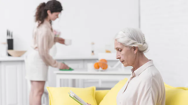 Haushaltshilfe wischt den Tisch, während eine Seniorin auf dem Sofa liest