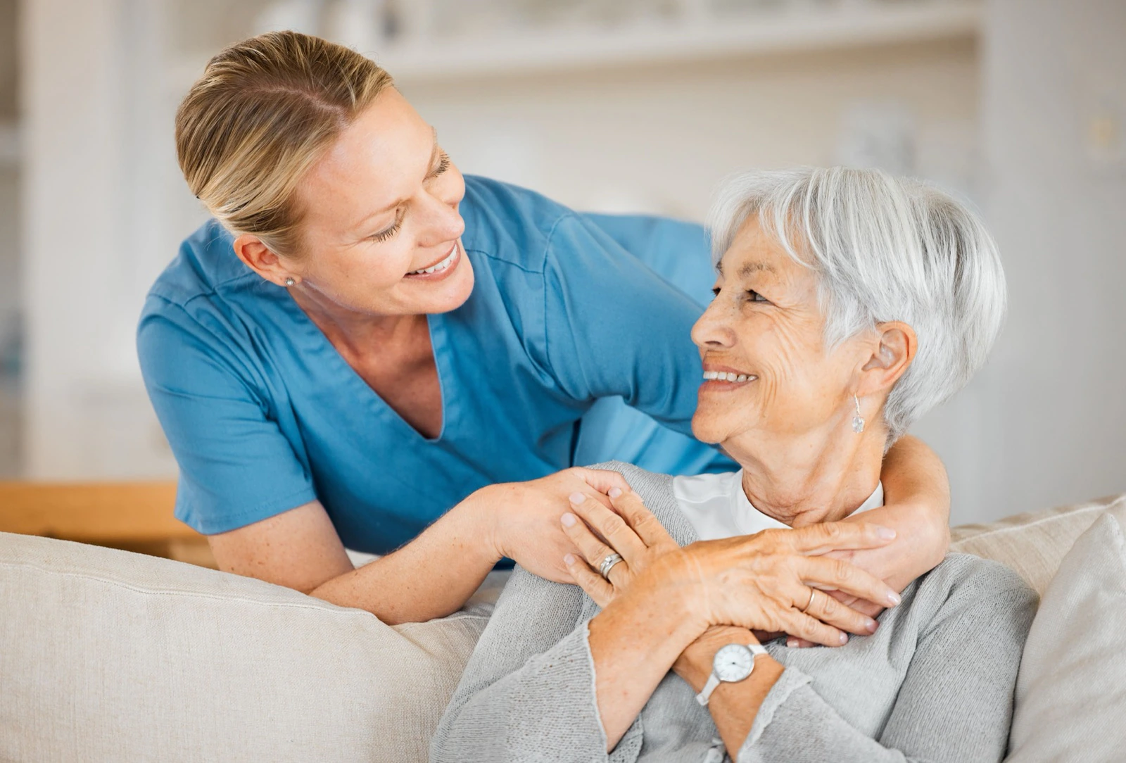 Pflegerin in blauer Dienstkleidung umarmt eine lächelnde Seniorin auf dem Sofa